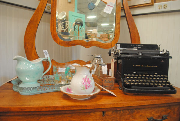 The photo features a black typewriter along with various vintage glass items sitting on a dresser. 